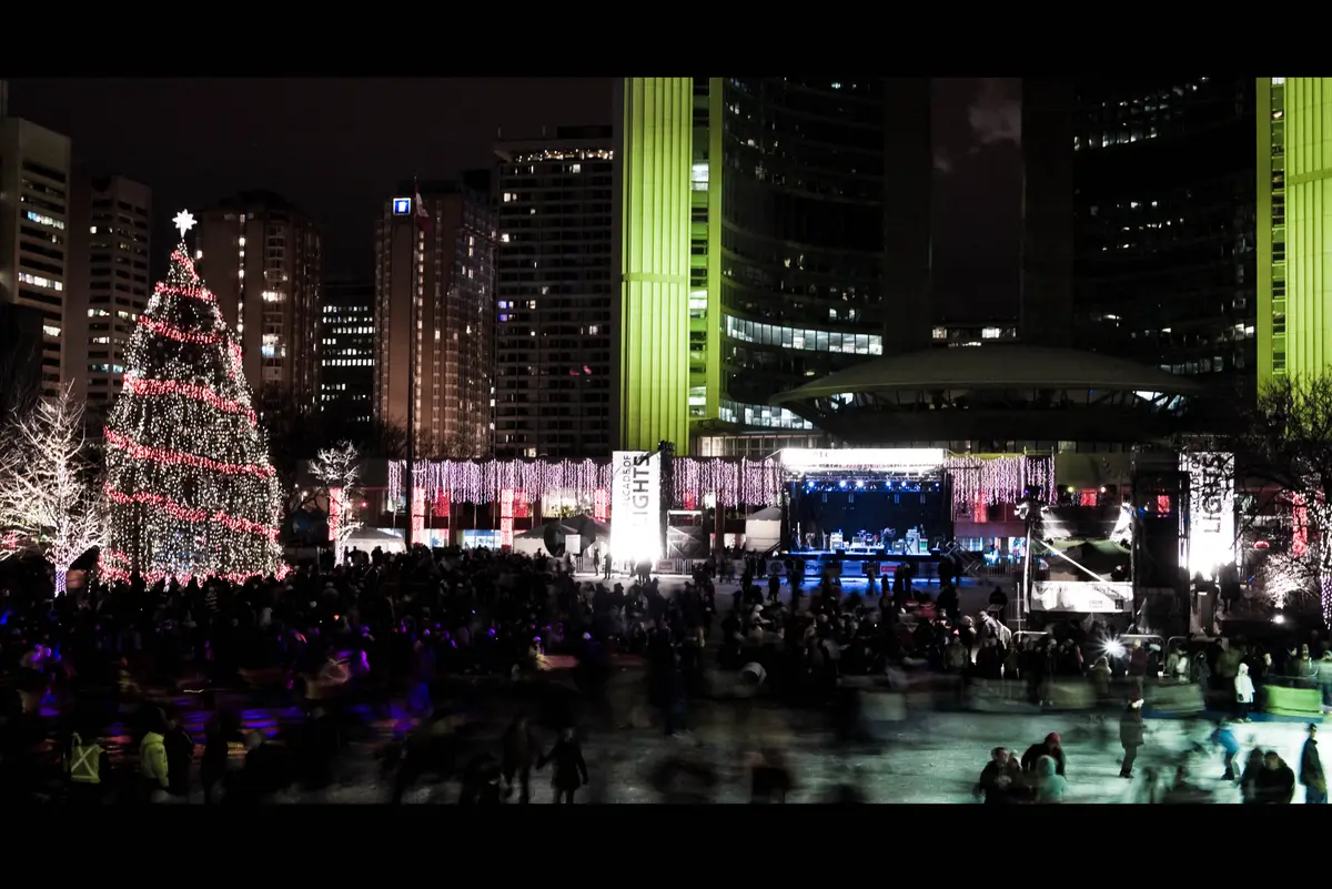A crowd gathered at night in Nathan Phillips Square beside a tall lit Christmas tree and a lit stage