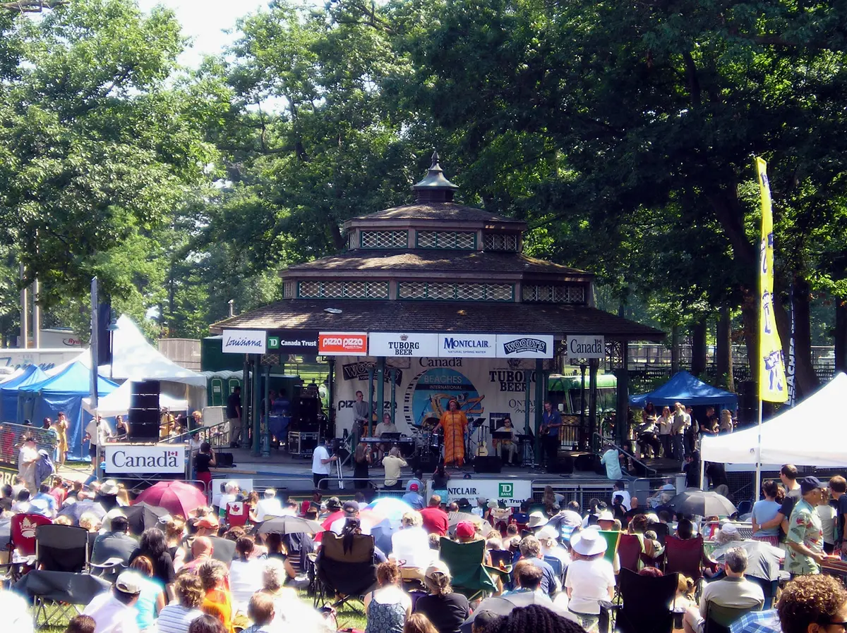 Crowd on the lawn in front of the Kew Beach grandstand, with a band on stage under the trees and banners along the front