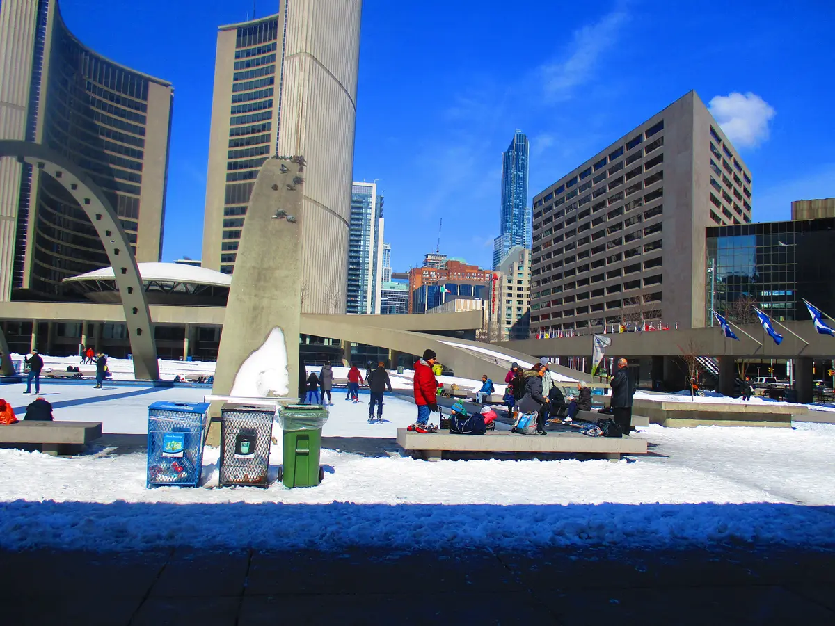 Skaters on an outdoor rink with snow along the edges, the City Hall towers behind and people seated on a plaza bench