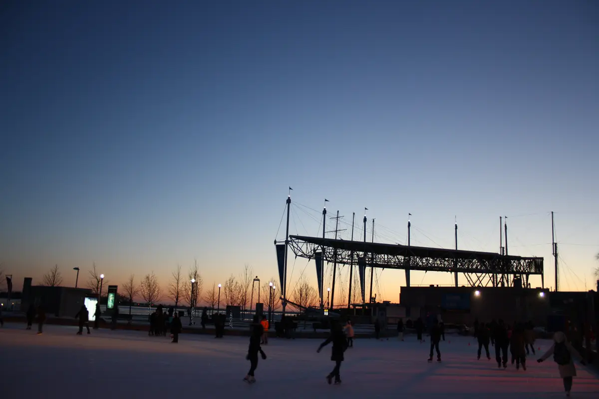 Skaters on an outdoor rink at dusk with an orange and blue sky behind a waterfront canopy